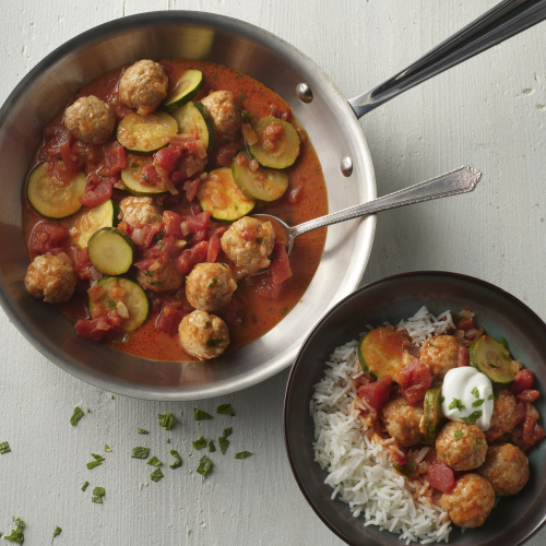 Chicken meatballs, sliced zucchinis, and diced tomatoes and onions simmer inside a curried red sauce in a silver pan while a plate of white long grain rice is topped by a scoop of the sauced entree with a dollop of sour cream.