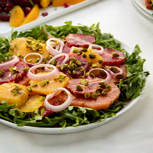 A colorful citrus salad featuring sliced oranges and grapefruits on a bed of arugula, garnished with red onion rings and chopped pistachios.