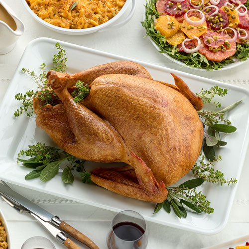 Roasted turkey garnished with herbs on a white serving platter, accompanied by side dishes including a citrus and onion salad and a bowl of mashed vegetables.