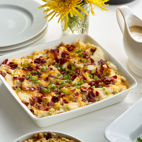 A dish of creamy mashed potatoes topped with crispy turkey bacon bits and sliced green onions, served on a white table with a gravy boat, plates, and a vase with yellow flowers in the background.