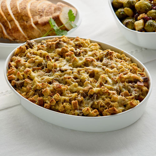 A ceramic dish filled with baked stuffing next to sliced bread and a bowl of cooked Brussels sprouts.